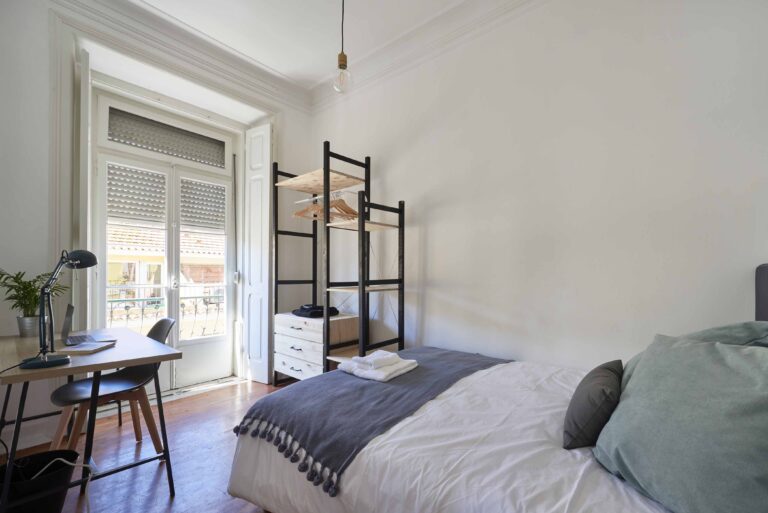 Minimalistic bedroom with a bed, desk, open wardrobe, and large windows showing rooftops.