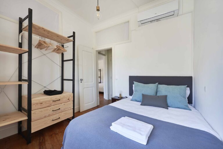 Minimalist bedroom with a double bed, blue blanket, shelves, drawers, and air conditioning.
