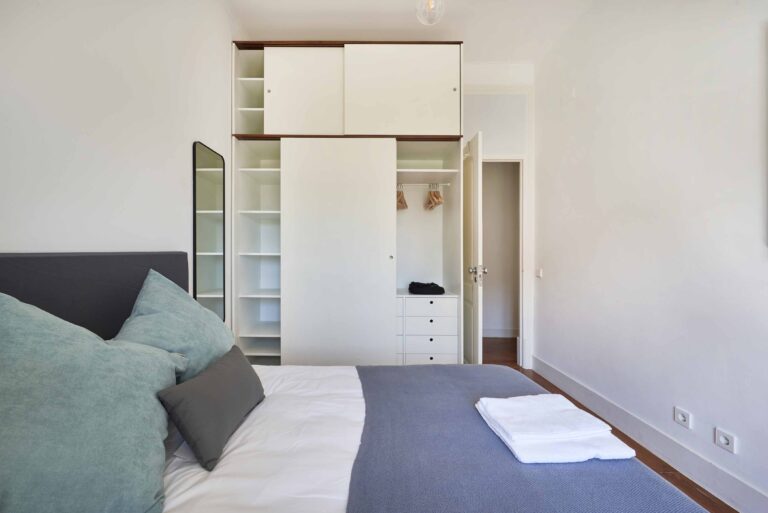 Bedroom with bed, pillows, blanket, open wardrobe, mirror, towels, and a door to another room.