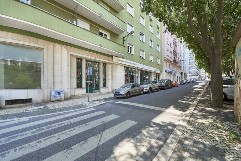 Street view of a green building with businesses below, parked cars, trees, and crosswalk nearby.