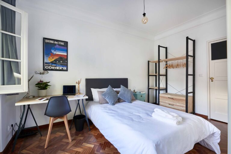 Modern bedroom with a bed, desk, laptop, plant, poster, open wardrobe, and window curtains.