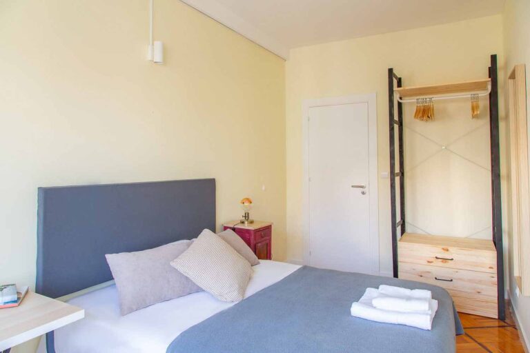 Simple bedroom with bed, red nightstand, lamp, open wardrobe, wooden hangers, and light walls.