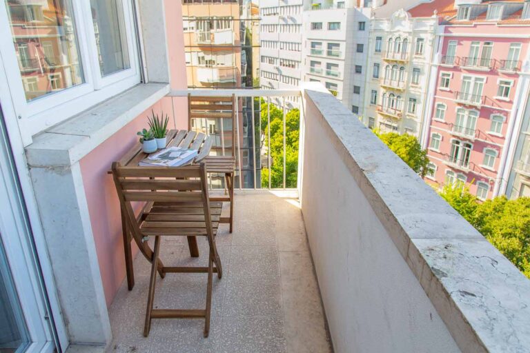 Small balcony with wooden table, chairs, potted plant; overlooks multistory buildings, clear sky.