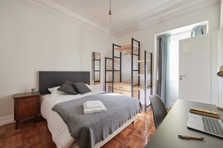 Minimalist bedroom with bed, nightstand, lamp, shelving, desk, laptop, mirror, door, window.