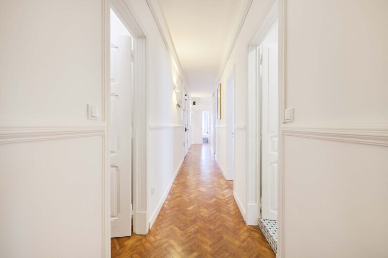 Long hallway with white walls, parquet floor, open doors, and natural light at the end.