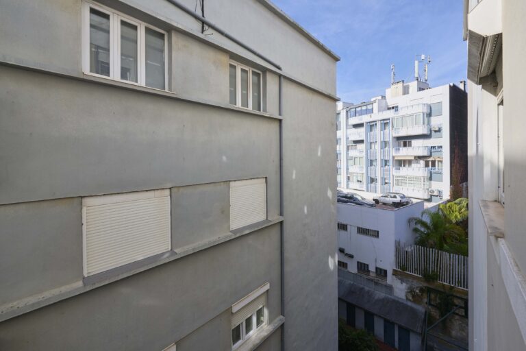 Urban scene with buildings, closed shutters, balconies, rooftop antennas, parked cars, and greenery.