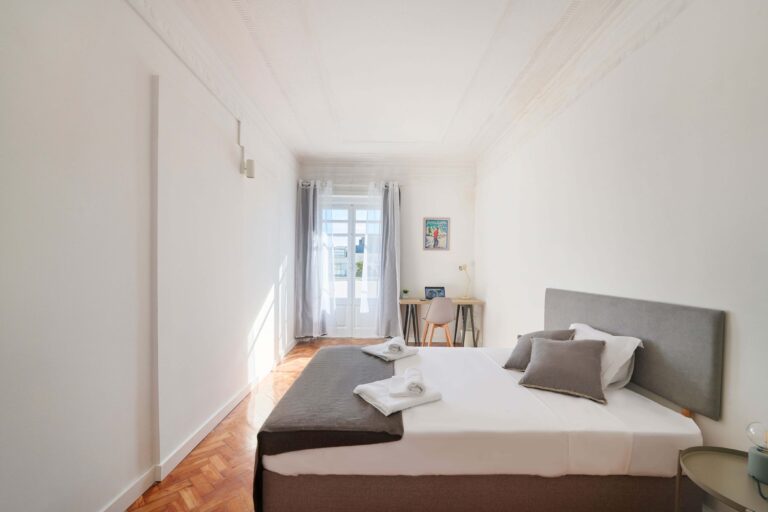 Bright room with double bed, gray headboard, desk, lamp, and natural light from a curtained window.