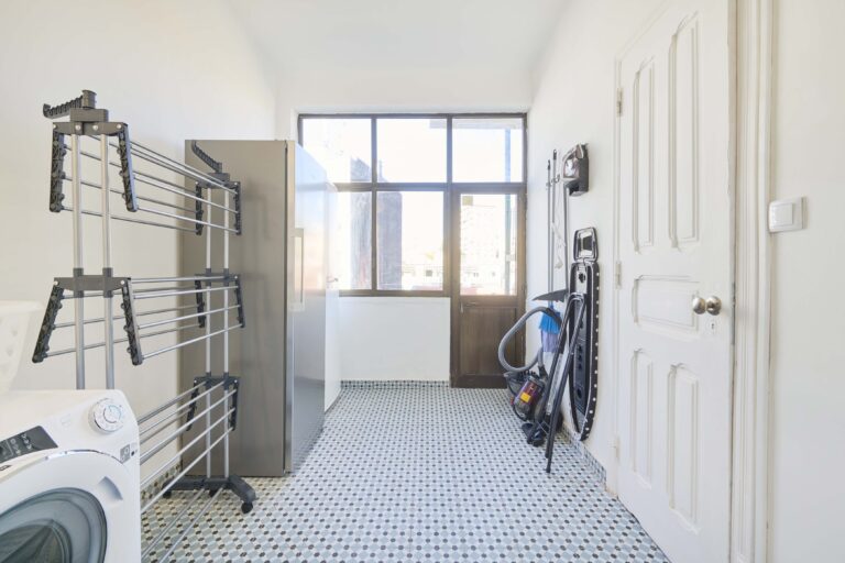 Utility room with washing machine, drying racks, vacuum, and ironing board on tiled floor.