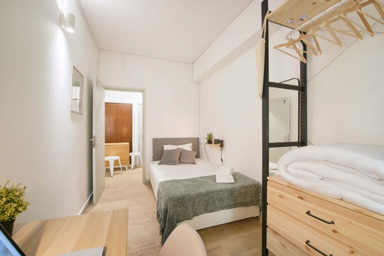 Small bedroom with single bed, wooden dresser, clothing rack, desk, chair, and minimalist decor.