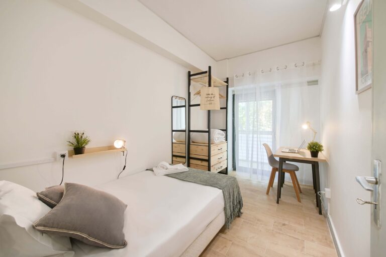 Minimalist bedroom with single bed, desk, shelves, lamp, plant, mirror, and sheer curtains.